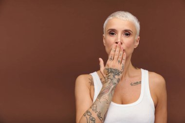 Beyaz yelekli, ağzı ağızla kaplayan ve hava öpücüğü gönderirken kollarını dövmelerle gösteren erkek benzeri bir kadın.