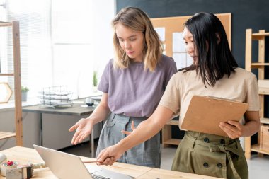 İki genç kadın stüdyoda masanın başında otururken, bir tanesi dizüstü bilgisayarı işaret ediyor ve meslektaşı da online bilgi paylaşımı yapıyor.