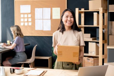 Çevrimiçi ofis çalışanlarının gülümseyen genç bayan çalışanı, paketlenmiş kutusuyla sana bakarken iş yerinde bilgisayarını kullanarak meslektaşına karşı ayakta duruyor.