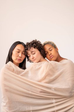 Asyalı, Kafkas ve Afrikalı kızlar, gözleri kapalı, beyaz pamuğa ya da şifon çarşafa sarılmış olarak birbirlerine yakın duruyorlar.