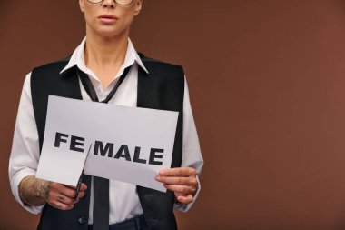 Zarif takım elbiseli, erkek görünümlü genç bir kadının orta kesimi kağıtta kadın kelimesinden heceyi keserek tecritte dururken erkekleşmesini sağlıyor.