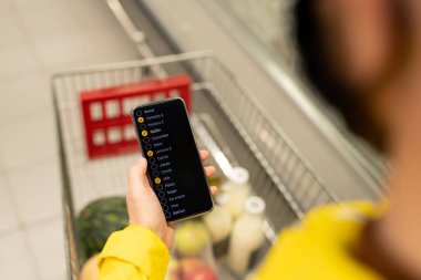 Markette kağıt torbalarla alışveriş arabalarını iterken akıllı telefondan alışveriş listesini karıştıran sıradan bir kadın alıcı.