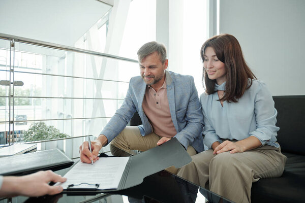 Happy middle aged couple signing document of selling and buying new car in auto center while sitting by table in front of camera