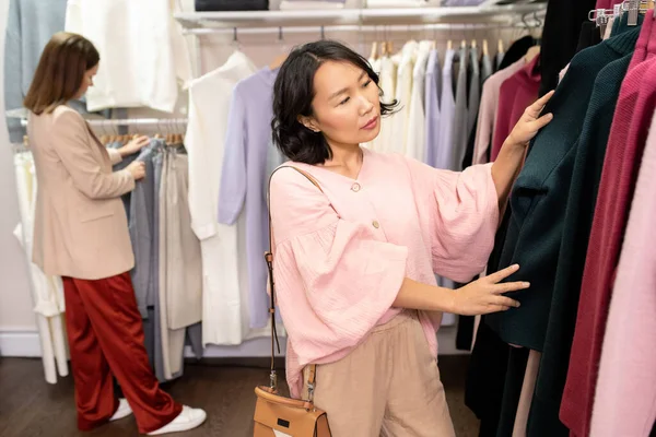 若いですアジアの女性の顧客でスマートカジュアルスタンドでラックで立っている新しいコレクションのセーターでブティックと自分自身のために1つを選択 — ストック写真