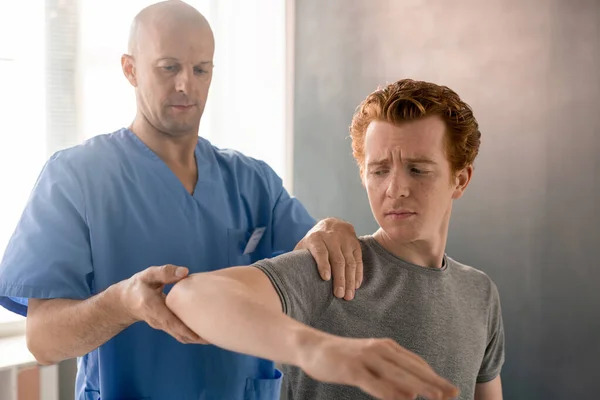 在现代康复诊所理疗训练中穿着蓝色制服支承弯曲手臂的成熟男性临床医生 — 图库照片