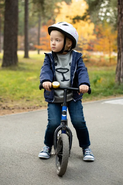 公園の松に対するカメラの前で彼のバランスの自転車によってアスファルトの道路に立っている犠牲者と安全ヘルメットで愛らしいブロンドの男の子 — ストック写真