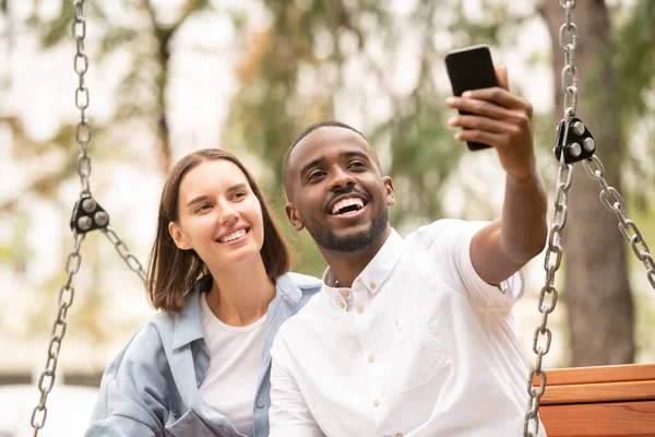 Młoda Wesoła Międzykulturowa Para Robi Selfie Siedząc Rano Ławce Parku — Zdjęcie stockowe