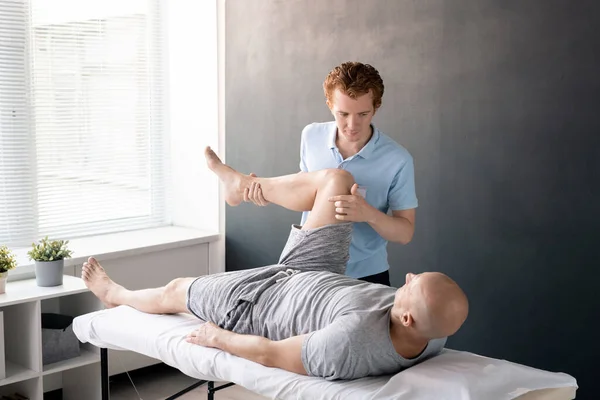 严肃的男性理疗师看着他的病人 腿弯在膝盖上 同时帮助他在康复中心进行一项体育锻炼 — 图库照片