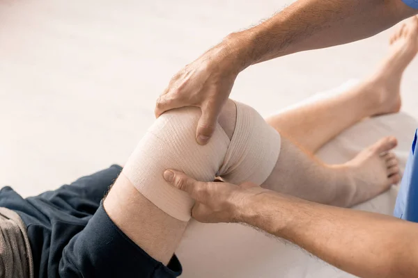 康复训练前在医疗过程中使用弹性绷带的残疾患者的男性临床医生双手包裹膝部 — 图库照片
