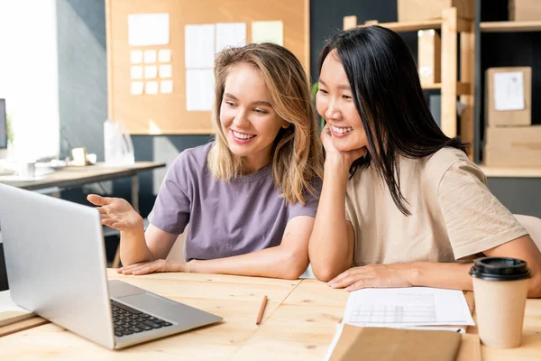 快乐的年轻亚洲女人坐在同事旁边 一边看笔记本电脑 一边讨论新的网上订货单 — 图库照片