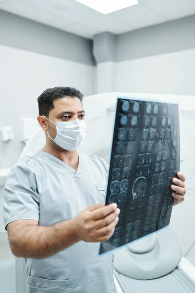 マスク中の深刻な成熟した混合レース放射線技師と医務室の環境に対する患者の頭のX線画像を分析する均一な — ストック写真