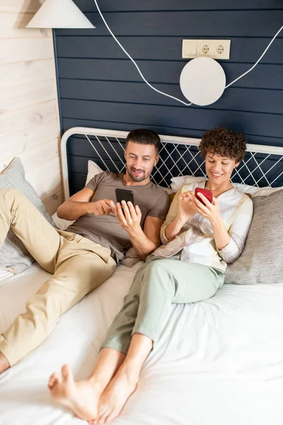 自宅環境でベッドでリラックスしながら 彼の妻好奇心オンラインビデオやソーシャルネットワークに投稿を示すスマートフォンを持つ若い男 — ストック写真