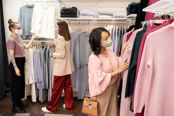 Young Asian female customer in mask and pink smart blouse standing by rack with new assortment of casualwear and looking through dresses