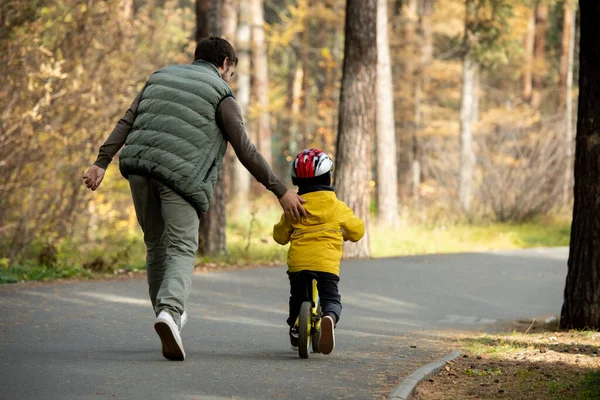 幼い息子をセーフティヘルメットとカジュアルウェアで走りながら 公園のバランス自転車の乗り方を教えていた父親の後ろ姿 — ストック写真
