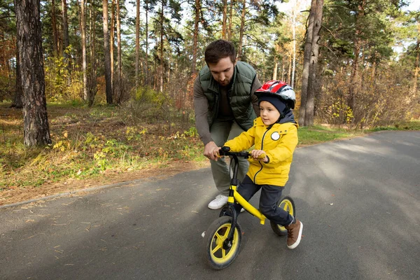 子供の父親は少し曲げながら 彼の小さな息子を助ける安全ヘルメットと犠牲者は公園のアスファルト道路に沿ってバランス自転車に乗るために着用 — ストック写真