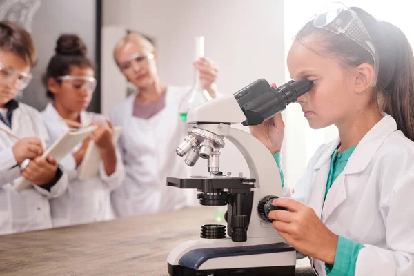 賢いです深刻な女子学生でホワイトコート見ますで顕微鏡によって机に対して彼女のクラスメートと先生作る化学実験 — ストック写真