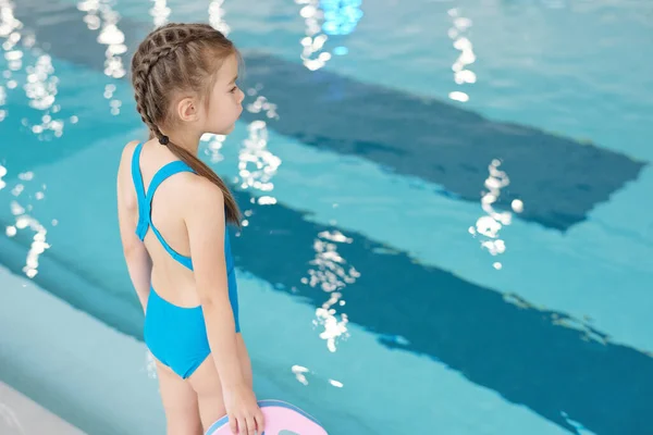在体育中心 身穿蓝色泳衣的小女孩站在摄像机前 看着游泳池中透明的水 站在游泳池边 看起来很可爱 — 图库照片