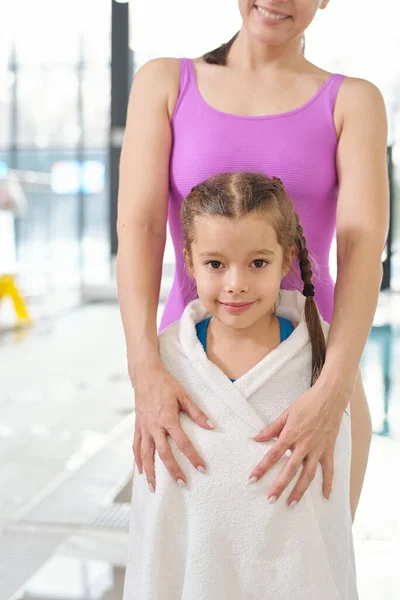 身着淡紫色泳衣的快乐年轻女性抱着湿透了的小女儿 裹着柔软的白色毛巾 站在镜头前 — 图库照片