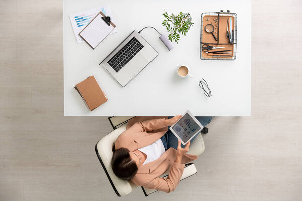 Overview of young elegant businesswoman with tablet sitting in white leather armchair by desk and analyzing financial charts and diagrams