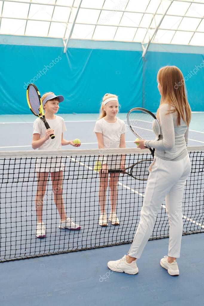 Two happy girls with tennis rackets standing by net in front of their instructor and listening to her explanations before training on stadium