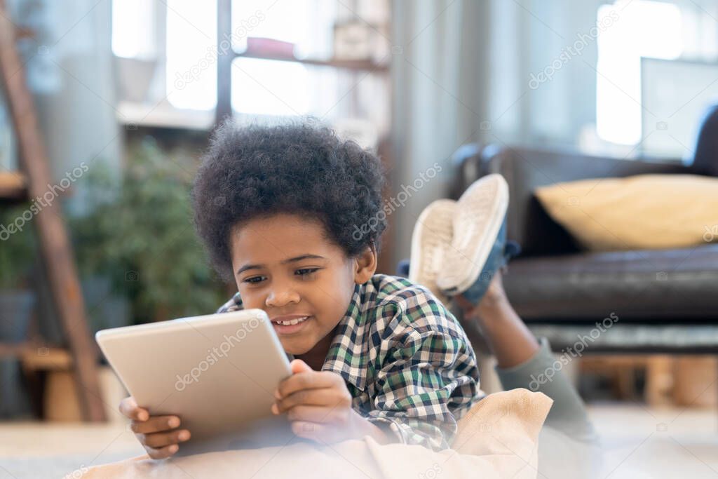 Happy elementary schoolboy with tablet lying on the floor by black leather couch in living-room and watching curious online video at leisure