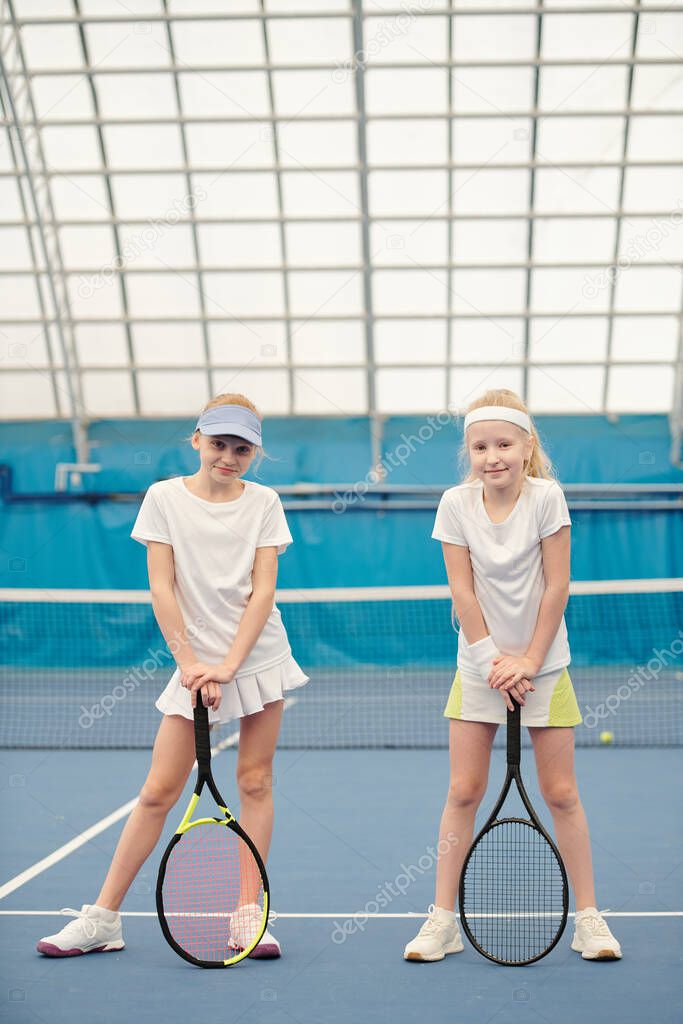 Two active teenage girls in white sportswear standing on tennis field at stadium against net and holding rackets ready for play or training