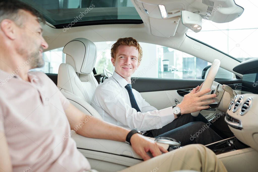 Happy young sales manager in formalwear sitting in new car and holding by steer during test drive with mature male client sitting next to him