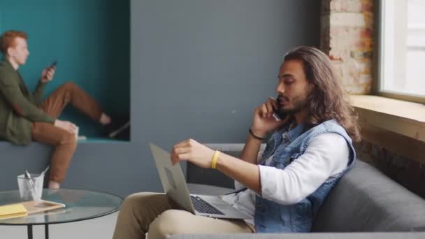 ノートパソコンの画面を見ながらソファに座って携帯電話で話す肩の長さの髪を持つハンサムな若いビジネスマンのPan 男の同僚は壁の床の間に座って電話を使って — ストック動画