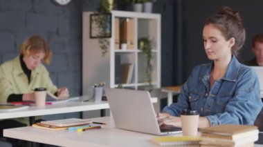 Kot ceketli genç bir iş kadınının ofiste otururken ve bilgisayarında kahve içerken çekilmiş PAN fotoğrafı.