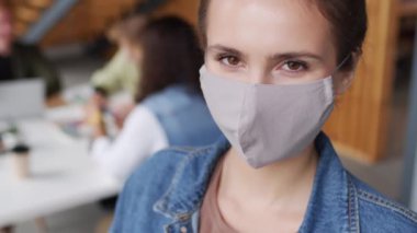 Kaşları çatık, maske takan ve ofisteki kameraya bakan genç iş kadınını yakından çek. Arka planda masanın etrafında oturan tanınmayan iş adamları