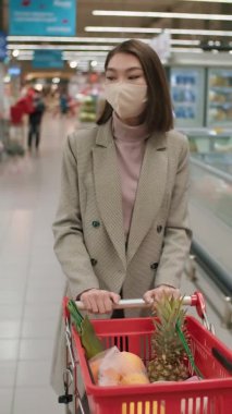 Maskeli, elinde alışveriş arabasıyla büyük bir süpermarketin koridorlarında yürüyen ve günlük yiyecek alışverişi yapan Asyalı bir kadının dikey orta pozu.