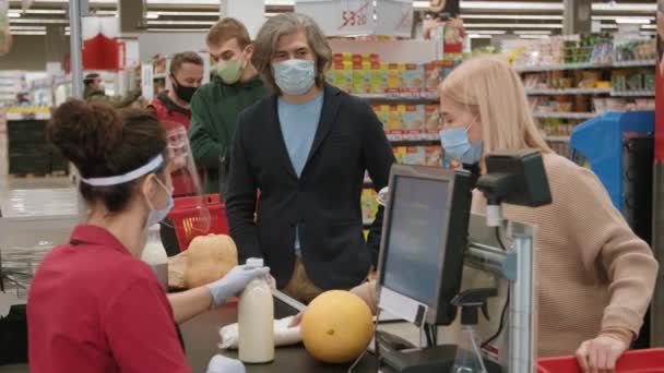 超级市场中带购物车和篮子站在队列中 戴着一次性口罩 而年轻女性出纳扫描产品的顾客的中等镜头 — 图库视频影像