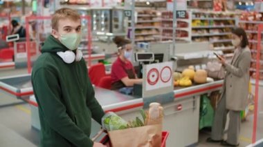 Yeşil kapüşonlu ve tek kullanımlık maskeli bir çocuğun orta boy portresi hipermarkette ürünlerle dolu bir alışveriş arabasıyla kasiyer ve kasiyerle kameraya bakıp gülümsüyor.