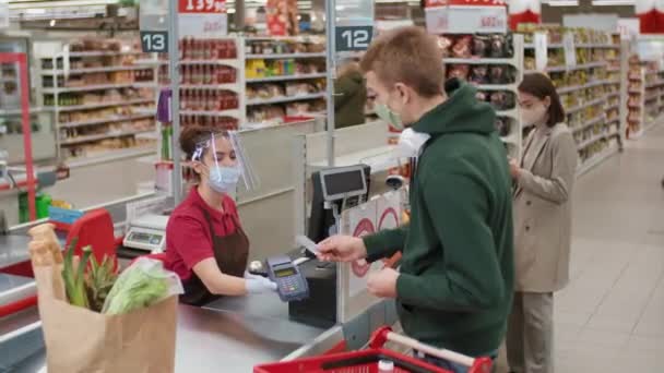 女性のレジが手渡した端末にクレジットカードで食品を支払う大きなスーパーマーケットでレジに立つ保護マスクの若い男の中出し — ストック動画