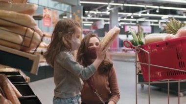 Koruyucu maskeli genç anne ve kızın, markette günlük yiyecek alışverişi yaparken orta yavaş çekimde taze ekmek seçerken ve alışveriş arabasına koyarken.