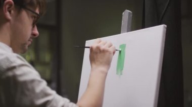 Elinde fırça olan konsantre erkek ressamın orta yavaş çekimde yakın çekimi karanlık boş sanat stüdyosunda beyaz tuvalde güzel bir şey yaratıyor.