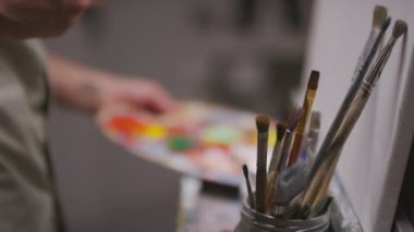 Orta bölüm, tanınmayan erkek sanatçının beyaz palette canlı renklerin yağlı boyalarıyla boya fırçasını karıştırarak karanlık, boş stüdyoda sanat eserleri yaratması.