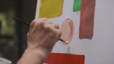 Renkli fırça resim çemberiyle tanınmayan erkek ressamın yakın çekimleri. Beyaz ve kırmızı yağlı boya boya tabloları, karanlık, boş bir sanat stüdyosunun tuvalinde.