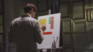 Arkaya bakan, boya fırçası ve yağlı boya paleti olan erkek ressamın karanlık sanat stüdyosundaki beyaz sehpanın önünde canlı renklerle geometrik şekiller çizdiği görüntüler.