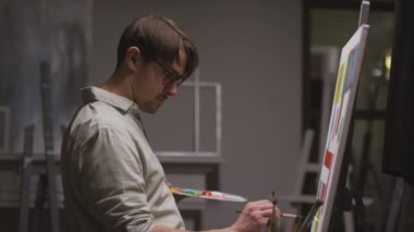 Palet ve el fırçasıyla konsantre olmuş yetenekli erkek ressamın orta yavaş görüntüsü..... karanlık sanat stüdyosunda tek başına duran beyaz sehpadaki yağlı boyaları resmedecek.
