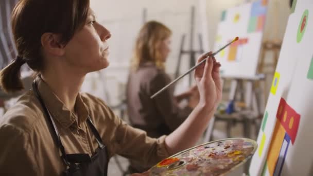 イーゼル ホールディング パレットと絵筆で白いキャンバスの前に座っている若い女性芸術家の中程度のクローズアップアートコースで抽象的な作品を作る — ストック動画