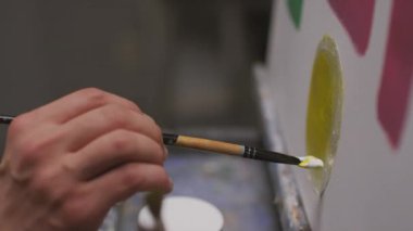 Tanımlanamayan ressamların yakın çekimi, sarı daire boyuyor, beyaz sehpada kalın vuruşlar yapıyor, karanlık, boş sanat stüdyosunda.