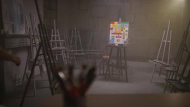 Tam yavaş çekimde bej gömlekli yetişkin bir sanatçının renkli geometrik şekiller resmiyle resim sehpasına bakarken karanlık odaya girmesi.