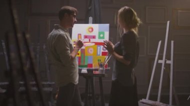 Ressamın yağlı boya paleti tuttuğu orta ölçekli arka plan görüntüleri ve onun kadın arkadaşı karanlık, boş bir sanat stüdyosunda resim tartışırken renkli geometrik şekiller tablosunun önünde duruyorlar.