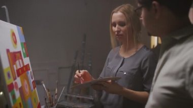 Kafkasyalı bir kadının, sıcak, karanlık sanat stüdyosunda erkek arkadaşıyla sanattan bahseden yağlı boyalarla parlak ve soyut bir tablo çizdiği orta boy bir yakın plan.