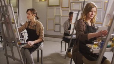 Tişört ve önlük giymiş üç genç kadının, parlak modern stüdyoda resim kursuna giderken tuval üzerinde oturup resim yapmasının tam karesi.