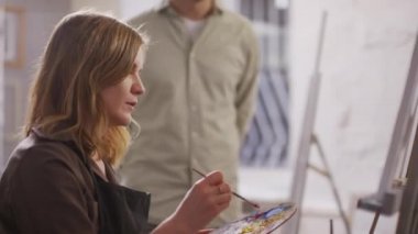 Renkli fırçayla tuvale resim yapan neşeli beyaz kadının orta boy yakın çekimi stüdyodaki sanat atölyesinde özel ders tavsiyelerini dinliyor.