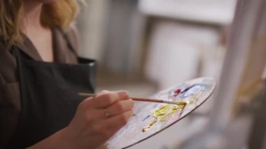 Kapalı alan sanat kursunda tuvale boya sürerek palette renkleri karıştıran genç bir kadının yakın plan çekimleri.