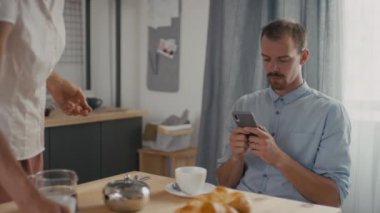 Kahvaltıda akıllı telefon kullanan cahil bir adamın orta yavaş çekimleri. Karısı masada duygusal kavgalar ve kavgalar ederken.