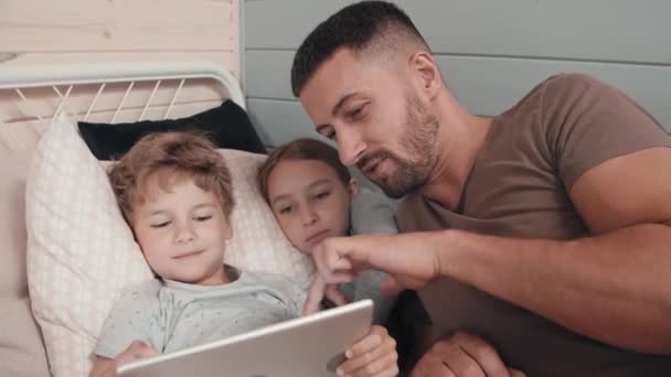 中等比例的胡子爸爸和可爱的小孩躺在床上孩子们的房间里玩平板电脑 — 图库视频影像